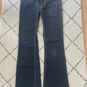 Mörkblå bootcut jeans från Roberto Cavalli - Säljer ett par mörkblå bootcut jeans från H&M x Roberto Cavalli med kontrastsömmar och broderad detalj på bakfickan. Jeansen har låg midja och snygg design.