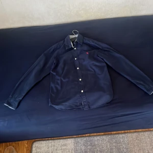  En skjorta, mörk blå  - En skjorta mörk blå med klassisk krage och vita knappar framtill. Liten röd broderad logga på bröstet. Perfekt för dig som vill ha en stilren och enkel look.