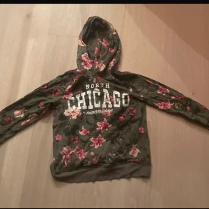 Grön blommig hoodie med tryck 'Chicago' - Säljer en grön hoodie med blommigt mönster och vit text 'North Chicago Awesome days' på bröstet. Tröjan har huva och långa ärmar, perfekt för en avslappnad stil. Blommorna är i rosa och röda nyanser.