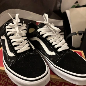 Svarta och vita sneakers från Vans - Klassiska Vans sneakers i svart med vita detaljer och vit sula. Skorna har snörning och den ikoniska vågformen på sidan. Perfekta för dig som gillar en tidlös och streetinspirerad stil.