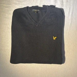 Marin blå v-ringad tröja från Lyle & Scott - Klassisk marin blå  v-ringad tröja från Lyle & Scott med gul broderad logga på bröstet. Tröjan är i mjukt material och har långa ärmar samt ribbade muddar vid ärmslut och nederkant. Perfekt för en stilren och enkel look.