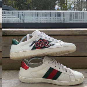 Gucci ace - Säljer ett par vita Gucci sneakers med broderad orm och röd/grön rand på sidan. Skorna har snörning, rund tå och platt sula. Bakpartiet har metallicdetaljer i grönt och rött. Perfekta för dig som vill sticka ut med lyxig streetstyle.
