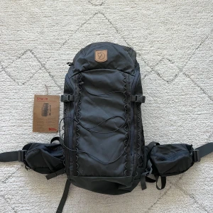 Svart ryggsäck från Fjällräven Singi 28 - Säljer en svart ryggsäck från Fjällräven, modell Singi 28. Ryggsäcken har flera fack, justerbara remmar och smarta snörningsdetaljer på framsidan. Märkets klassiska logga i läder syns upptill. Perfekt för äventyr och vardag.