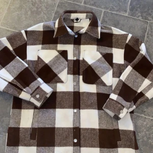 Brun och vit rutig jacka från H&M - Säljer en brun och vit rutig overshirt från H&M. Skjortan har stora bröstfickor med lock, knappar framtill och klassisk krage. 
