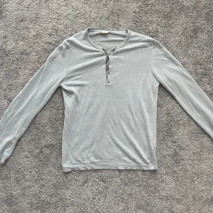 Ljusgrå långärmad tröja från Hugo Boss - Säljer en stilren ljusgrå långärmad tröja från Boss Orange. Tröjan har knappar framtill och är tillverkad i ett mjukt, stickat material. Perfekt för lager-på-lager och enkel att matcha med olika outfits.