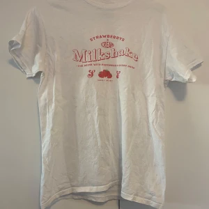 Vit t-shirt med milkshake-tryck från Heartbreak - Vit t-shirt från Heartbreak med rött tryck på bröstet där det står 'Strawberrys & Cream Milkshake'. T-shirten är i 100% bomull och har en klassisk passform med korta ärmar. Perfekt för dig som gillar retroinspirerade prints och mjuka material.
