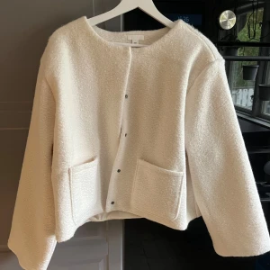 Vit teddyjacka med fickor - Säljer en vit teddyjacka med rund halsringning och stora framfickor. Jackan har tryckknappar framtill och är croppad i modellen. Perfekt för dig som gillar en mysig och trendig stil.