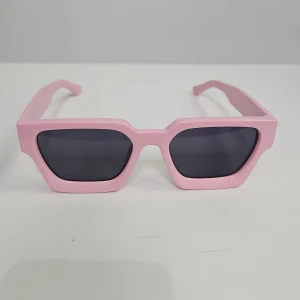 Rosa kantiga solglasögon - Coola solglasögon med bred, kantig båge i ljusrosa plast och mörka glas. Perfekt accessoar för dig som vill sticka ut med en trendig och lekfull stil.