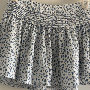 Vit blommig kortkjol med blått mönster - Supersöt vit kortkjol med småblommigt blått mönster och smockad midja. Kjolen har volang och är lätt utställd, perfekt för varma dagar. Materialet känns mjukt och luftigt, vilket gör den bekväm att bära.