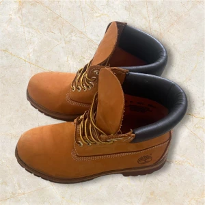 Bruna boots från Timberland - Klassiska bruna boots från Timberland med robust snörning och svart vadderad kant runt skaftet. Skorna har grov sula och är perfekta för höst och vinter. Ikonisk logga på sidan och slitstarkt material.