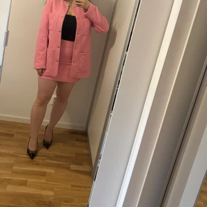 Rosa tweed kavaj och kjol-set - Säljer helt ny  set med kavaj och matchande kjol. Kavajen har guldknappar och stora fickor framtill, och kjolen är kort med två fickor. Materialet är strukturerat och känns mjukt. Perfekt för dig som vill sticka ut med färg!