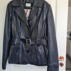 Svart skinnjacka med bälte från Tempo & Tappa - Snygg svart skinnjacka med croco-mönster från Tempo & Tappa. Jackan har bälte i midjan, knappar framtill och långa ärmar. Fodret har ett unikt tryck. Perfekt för dig som vill sticka ut med en cool look.
