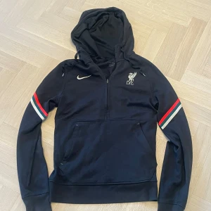Svart Nike Liverpool hoodie med röd och vit detalj - Svart hoodie från Nike med Liverpool FC-logga broderad på bröstet. Tröjan har huva, halv dragkedja och röda samt vita ränder på ärmarna. Tillverkad i Dri-FIT-material som andas. Perfekt för fotbollsälskare! Storlek XS