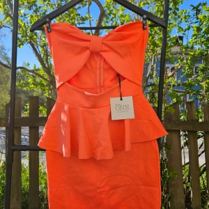 Orange axelbandslös klänning med rosett  - Säljer en orange axelbandslös klänning med stor rosettdetalj framtill och peplumvolang i midjan. Klänningen har dragkedja bak och figurnära passform. Perfekt för dig som vill sticka ut!