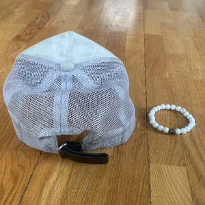 Ljusgrå keps och vitt armband från Rovior. Bra skick. - Säljer en ljusgrå keps från Rovior med mesh-tyg på sidorna och baksidan samt justerbart spänne. Framtill finns en patch med ett svartvitt motiv av Eiffeltornet. Perfekt accessoar för en stilren look. Kepsen kommer med ett vit armband.