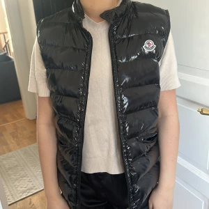 Svart dunväst från Moncler - Snygg svart glansig dunväst från Moncler med klassisk logga på bröstet. Västen har ståkrage och quiltad design, perfekt att slänga över en tröja för en cool look. Innerfoder med märkning 'LONGUE SAISON'.