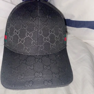 Svart keps med monogram - Säljer en svart keps från med klassiskt GG-monogram över hela kepsen. På sidorna finns gröna och röda ränder. Justerbar baktill och fodrad med svart tyg. Perfekt för dig som gillar exklusiva accessoarer.