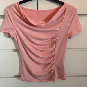 Ljusrosa topp med drapering - Säljer en ljusrosa topp med korta ärmar och snygg drapering framtill. Toppen har en bred, något båtringad halsringning och figurnära passform. Perfekt för dig som vill ha en feminin och trendig look.