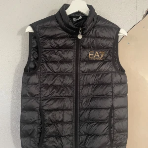 Svart väst och Tracksuit från EA7 Emporio Armani - Säljer en svart quiltad väst från EA7 Emporio Armani med dragkedja framtill och logga i guld på bröstet. Perfekt för lager-på-lager och har två fickor med dragkedja.                                                     800 kronor för västen och 600 kronor styck för jackan och byxorna. 