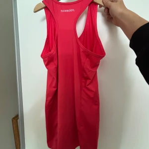 Röd träningstopp från Newbody - Säljer en röd ärmlös träningstopp från Newbody i funktionsmaterial. Modellen är tight och har brottarrygg, perfekt för gym eller löpning. Snygg och enkel design med rund halsringning.