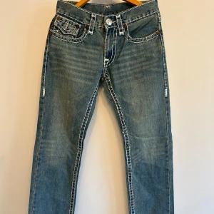 Blå jeans från True Religion med vita sömmar - Säljer ett par klassiska blå jeans från True Religion med tydliga vita kontrastsömmar och snygga fickdetaljer bak. Jeansen har normal passform och är perfekta för dig som gillar streetwear och vill sticka ut lite extra.💞 { Längd: 103,5 cm, Innerbenslängd: 78,5 cm}