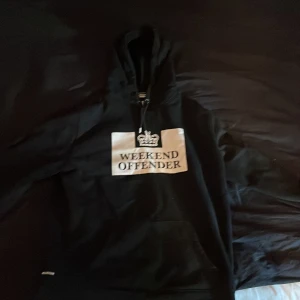 Svart hoodie från Weekend Offender - Svart hoodie från Weekend Offender med vit logga och text på bröstet. Klassisk huva med dragsko och magficka framtill. Perfekt för en avslappnad stil.