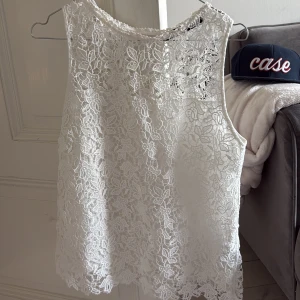 Vit spetslinne med blommönster - Säljer ett vitt ärmlöst linne med vackert blommigt spetsmönster. Perfekt för varma dagar och ger en romantisk känsla till outfiten. Linnet har rund halsringning och fina detaljer längs nederkanten.