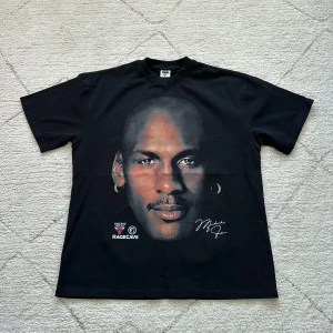 Svart t-shirt med Michael Jordan-tryck från RageCave - Svart t-shirt med stort porträtt av Michael Jordan på framsidan och ikonisk basketbild med texten 'RageCave Wont Miss' på baksidan. Rund hals och normal passform. Perfekt för basketfans!