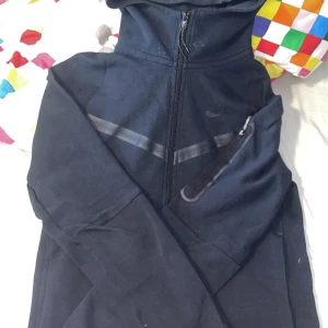 Svart zip-hoodie från Nike - Säljer en svart hoodie från Nike med hel dragkedja och huva. Jackan har en diskret logga på bröstet och en snedställd bröstficka med dragkedja. Perfekt för en sportig look och enkel att matcha.