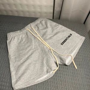Grå shorts från Essentials Fear of God - Snygga ljusgrå shorts från Essentials Fear of God med svart logga på ena benet. De har resår i midjan och långa snören framtill för justering. Perfekta för en avslappnad stil.