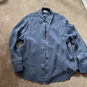 Blå skjorta från & Other Stories - Säljer en stilren blå skjorta från & Other Stories. Skjortan har klassisk krage, knäppning framtill och långa ärmar. Perfekt för dig som gillar en enkel men snygg look. Materialet känns mjukt och har en lätt glans.