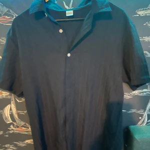 Mörkblå kortärmad skjorta med knappar och tillhörande kortbyxor - Säljer ett sommar sätt av en stilren mörkblå skjorta med korta ärmar och vita knappar samt ett par mörkblåa kortbyxor. De är tillverkade i ett lätt material som passar perfekt till varma dagar. Enkel och snygg design som funkar till många tillfällen. Säljes då jag fick de i present men de inte passade min stil. Även osäker vad de är för märke.