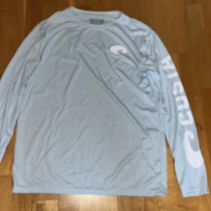 Ljusblå långärmad tröja från Costa - Säljer en ljusblå långärmad tröja från Costa med vit logga på bröstet och stort tryck på ena ärmen. Tröjan har rund halsringning och är tillverkad i ett tunt, syntetiskt material som passar perfekt för aktiva dagar. 