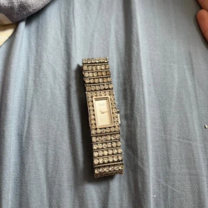 Glittrig silverklocka från D&G - Säljer en glittrig klocka från D&G i silverfärg med rektangulär urtavla och bred länkarmband täckt av gnistrande stenar. Klockan har en exklusiv känsla och D&G-logga på både urtavla och spänne. Perfekt accessoar för att sticka ut. Men fungerar inte tyvärr 