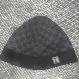 Svart  mössa från Louis Vuitton - Säljer en mörkgrå mössa från Louis Vuitton med diskret rutigt mönster och svart uppvikt kant. LV-logga framtill. Perfekt accessoar för kyliga dagar.