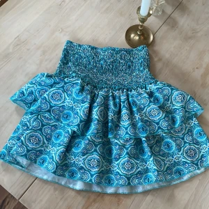 Blå mönstrad kortkjol med volang - Supersöt blå kortkjol med vitt och turkost mönster. Kjolen har bred smock i midjan och volang nertill som ger en luftig känsla. Perfekt för varma dagar och enkel att styla med en enkel topp.