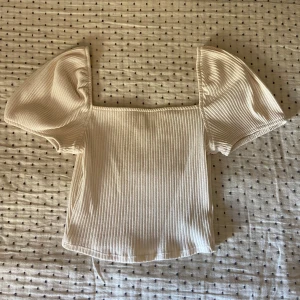 Beige ribbad topp med puffärmar - Säljer en beige ribbad topp med fyrkantig ringning och korta puffärmar. Toppen har en croppad passform. Toppen har knappt använts och är i mycket bra skick.