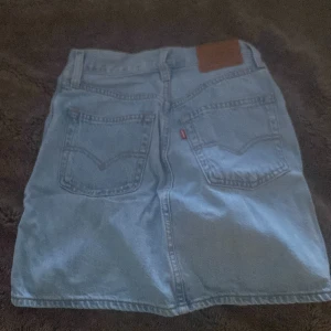 Jeanskjol med knappar från Levi's - Säljer en klassisk ljusblå jeanskjol från Levi's med knappar framtill och två bakfickor. Kjolen har bälteshällor och en rak passform som passar perfekt till vardags. Ikonisk Levi's-lapp baktill i midjan.