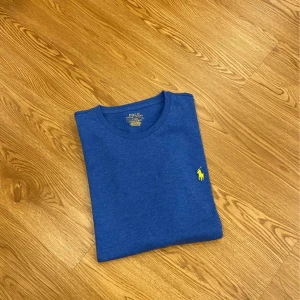 Blå t-shirt från Polo Ralph Lauren - Snygg blå t-shirt från Polo Ralph Lauren med klassisk rund hals och gul broderad logga på bröstet. Perfekt basic-plagg med normal passform och korta ärmar. Tillverkad i mjuk bomull för maximal komfort.