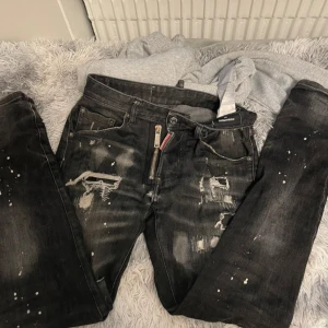 Svarta jeans med slitningar från Dsquared2 - Säljer ett par svarta jeans från Dsquared2 med coola slitningar och färgstänk. Jeansen har dragkedja och klassisk femficksdesign. Märkeslapp bak i midjan och snyggt distressed look.
