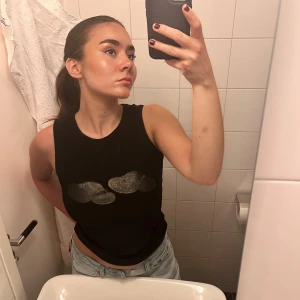 Svart ärmlös topp med glittriga moln - Svart ärmlös linnetopp från The Ragged Priest med två glittriga silvermoln framtill. Perfekt för dig som vill sticka ut med en unik detalj. Mjuk och stretchig känsla.