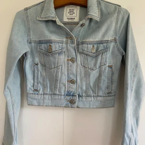 Ljusblå croppad jeansjacka från Pull&Bear - Säljer en ljusblå croppad jeansjacka från Pull&Bear. Jackan har klassisk krage, knappar framtill och två bröstfickor med lock. Perfekt för dig som gillar en trendig och avslappnad stil.