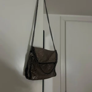 Glittrig axelväska med kedja - Säljer en glittrig axelväska i metallic-brun färg med svarta kedjedetaljer runt kanterna och som axelrem. Väskan har lock med tryckknapp och är perfekt för att lyfta din outfit.