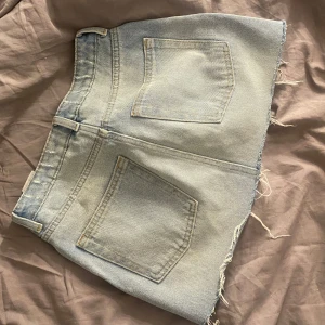 Ljusblå jeanskjol från H&M - Säljer en ljusblå jeanskjol från H&M i storlek 36. Den har råa, fransiga kanter och klassiska fem fickor. Perfekta för varma dagar och har en avslappnad look.