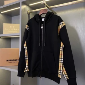 Svart Burberry Zip Hoodie - Svart hoodiejacka från Burberry med dragkedja och klassiska rutiga paneler längs ärmarna och sidorna. Jackan har huva med dragsko och två fickor framtill. Perfekt för en stilren och trendig look