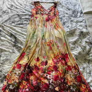 Blommig långklänning med smockad överdel - Färgglad långklänning med blommigt mönster i rosa, rött och gult. Klänningen har smockad överdel, v-ringning och är ärmlös. Perfekt för sommarens varma dagar. Storkleken är oklar men runt 40-50