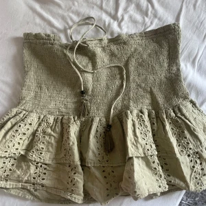 Beige kjol med volang och broderade detaljer - Supersöt beige kjol med smock upptill och två lager volang med broderade hålmönster. Kjolen har knytband i midjan med tofsar. Perfekt för en somrig och avslappnad look.
