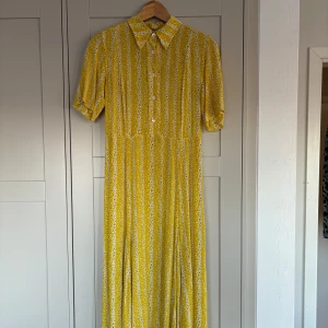 Gul blommig klänning från arket  - Charmig gul klänning med vita blommor och knappar framtill. Den har en klassisk krage och korta ärmar, perfekt för en solig dag. Klänningen är lång och ger en härlig sommarkänsla. Storlek 34/ xs