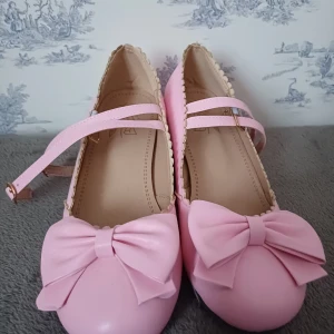 Rosa ballerinaskor med rosett - Söta rosa ballerinaskor med en stor rosett framtill och en liten klack. Skorna har en dekorativ kant och ett justerbart band över foten för extra stil. Perfekta för en lekfull och feminin look.