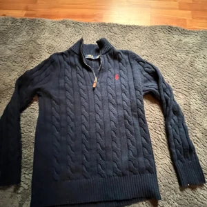 Mörkblå  stickad tröja från Ralph Lauren - Säljer en stilren mörkblå stickad tröja från Ralph Lauren med dragkedja och ribbad krage. Tröjan har ett diskret rött broderat emblem på bröstet och långa ärmar. Perfekt för kyliga dagar. Storleken är L🍾🍾